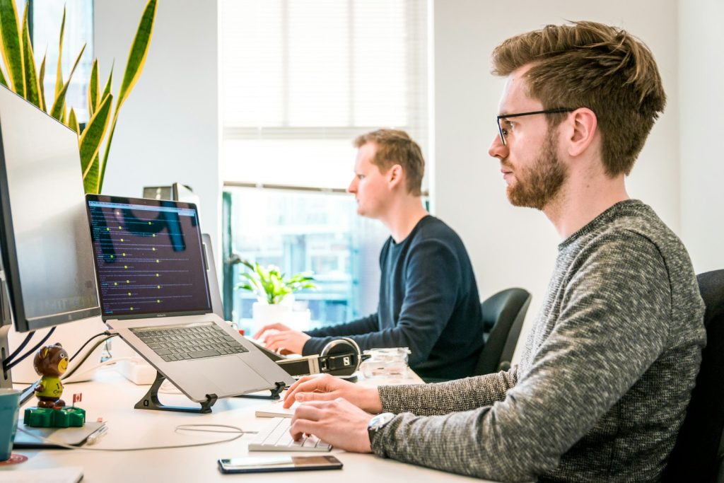 Two colleagues at a bright office desk working through a plan together on a laptop and a desktop monitor.