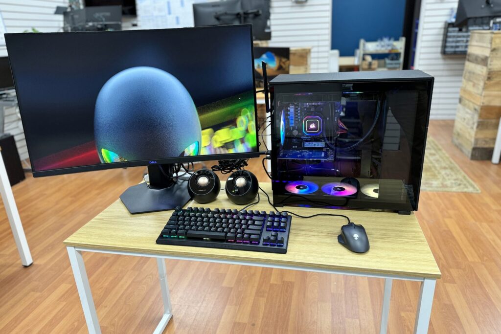 A custom built desktop PC with a glass side panel and lit cooling fans on a showroom table, beside a monitor, a backlit keyboard, a mouse and small speakers.