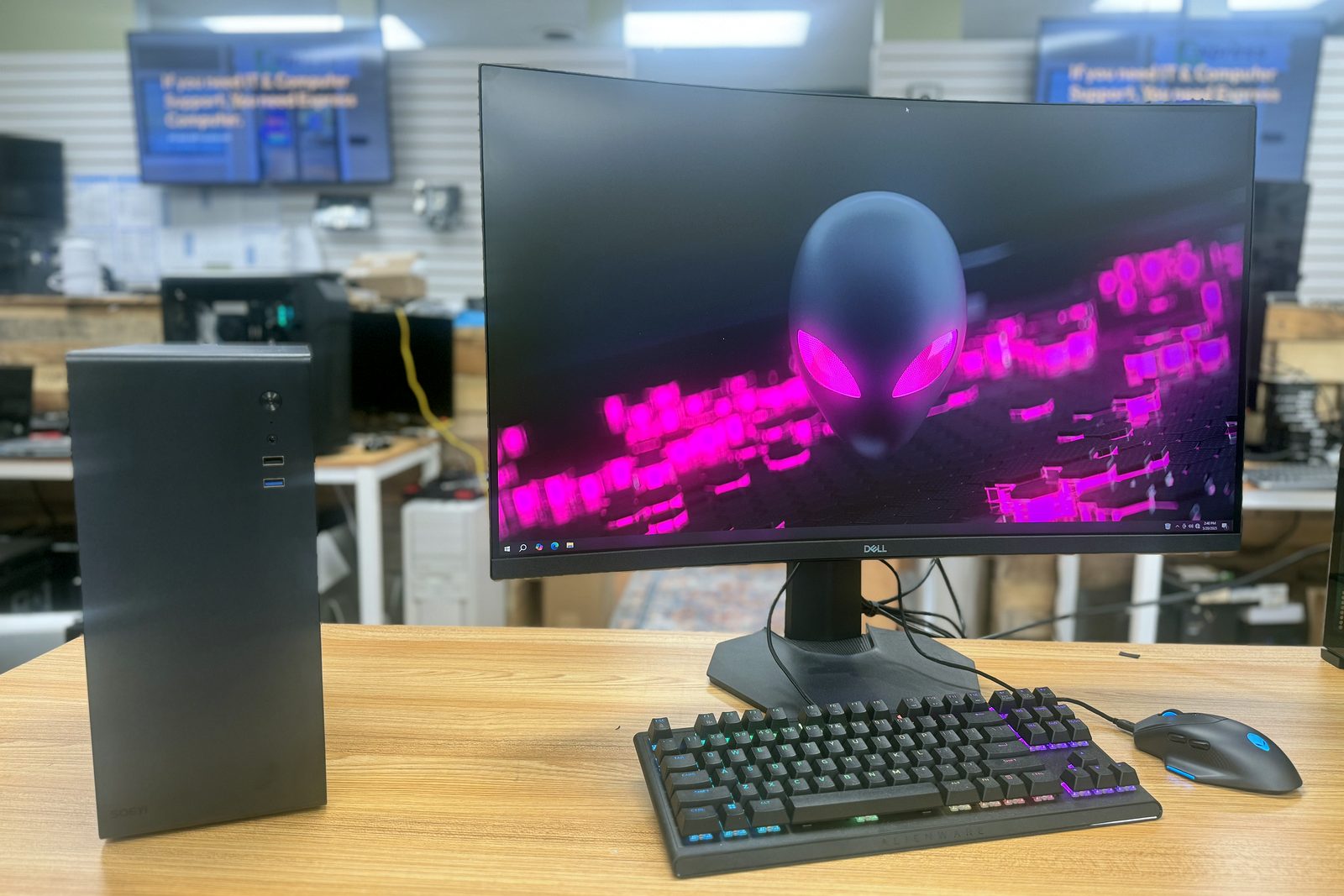 A black desktop tower beside a widescreen monitor, a backlit keyboard and a mouse on a showroom display table.