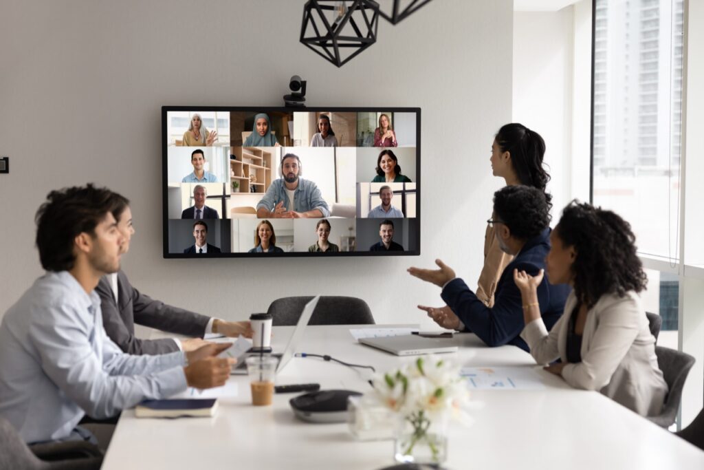 Team in a conference room on a video call with a wall-mounted screen showing twelve remote participants