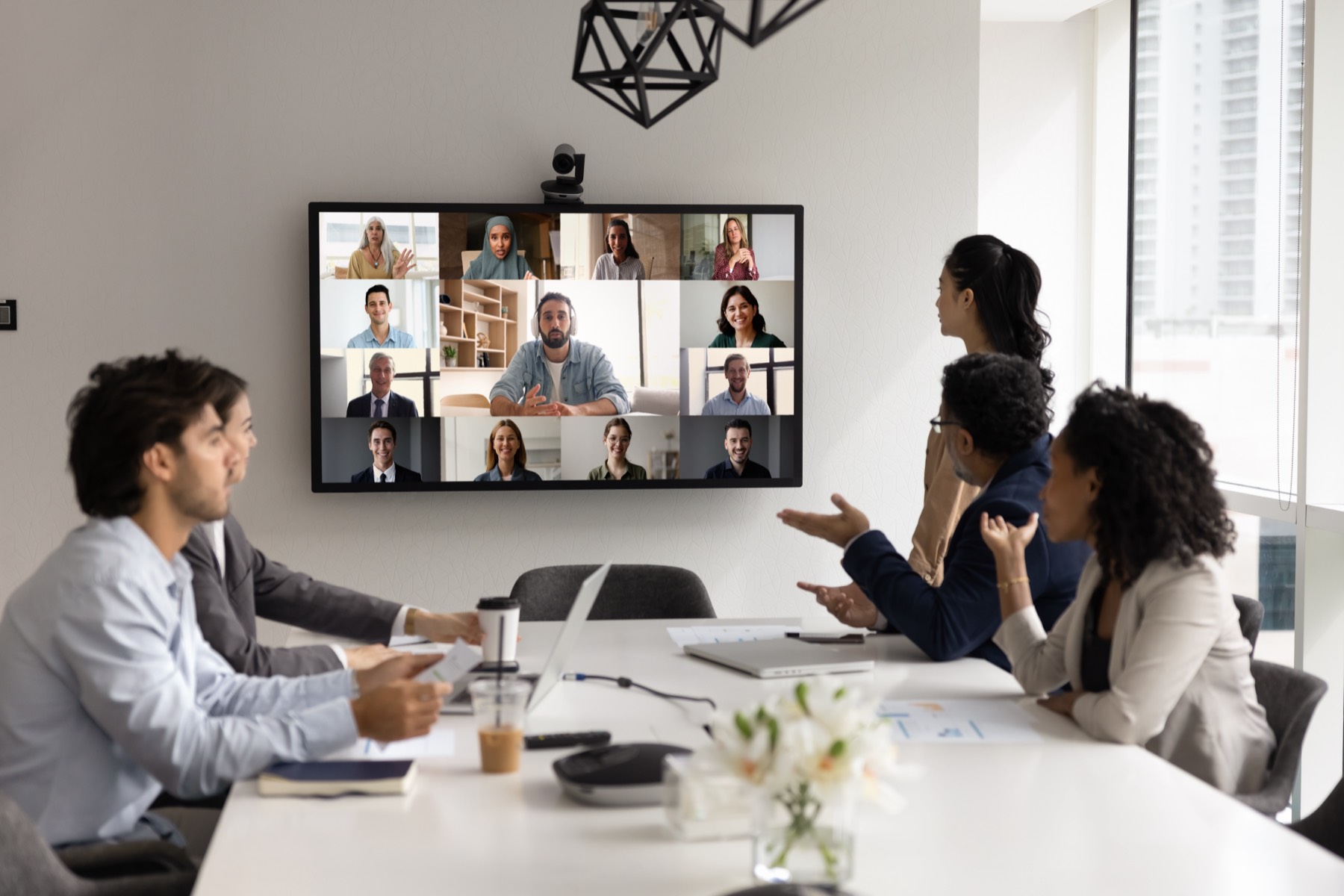 Team in a conference room on a video call with a wall-mounted screen showing twelve remote participants