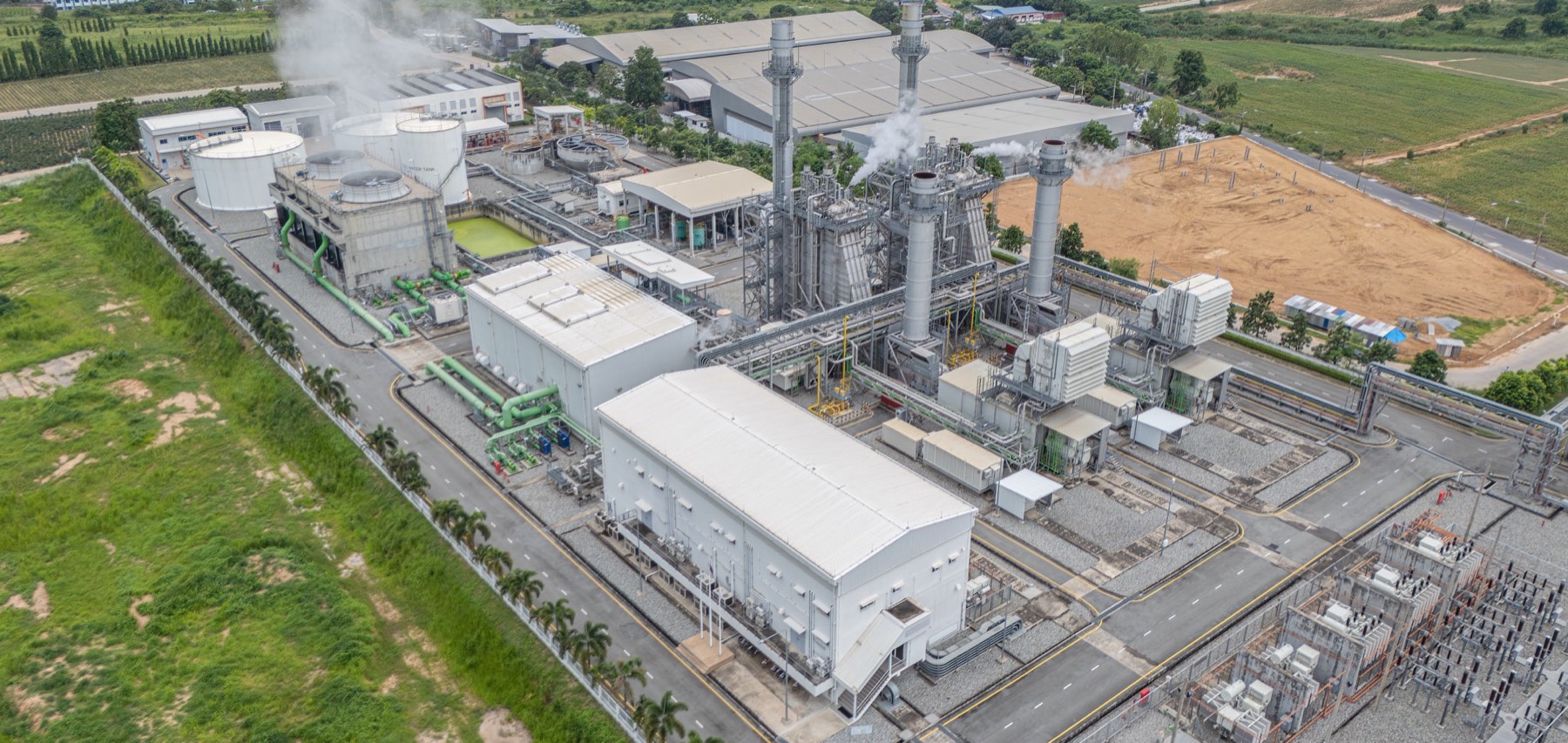 Aerial view of a combined cycle gas power plant with stacks, turbine halls, a cooling tower, storage tanks and a switchyard.