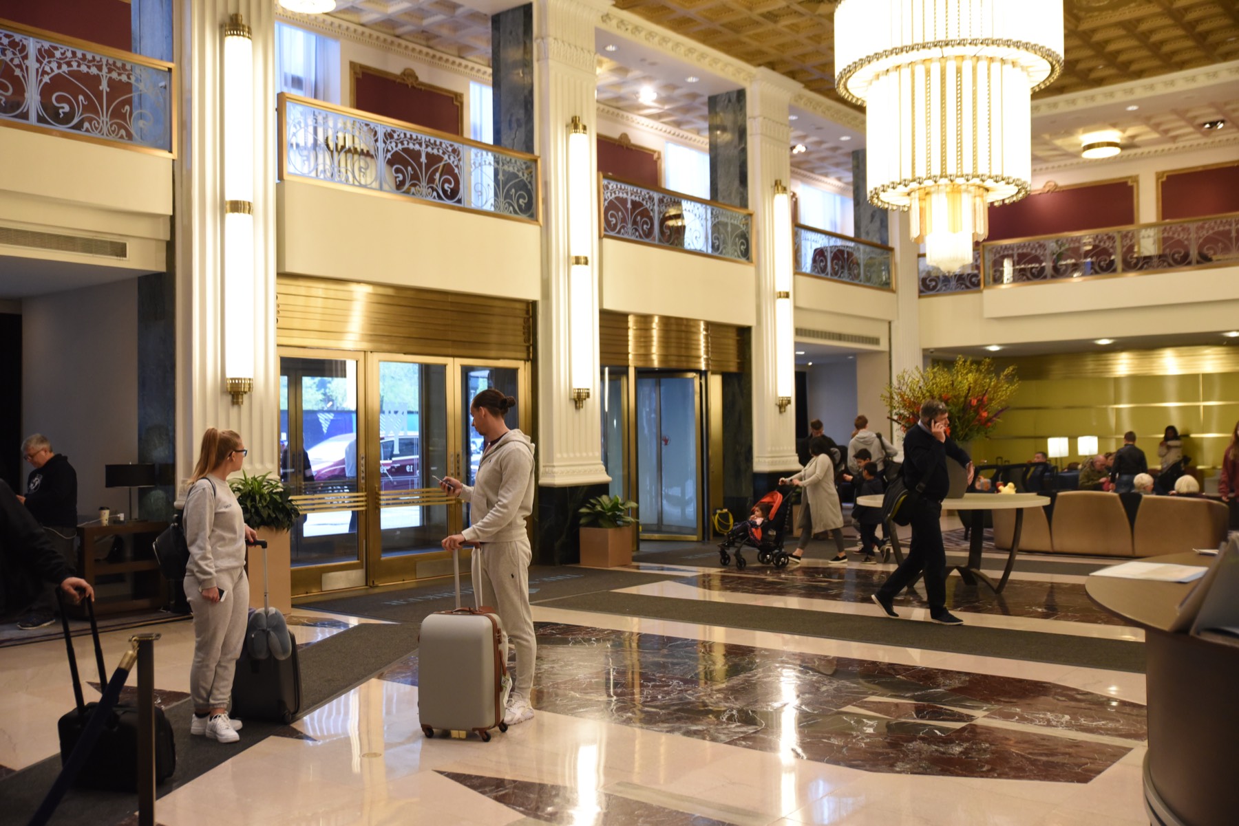 Guests with rolling luggage in a grand hotel lobby, with a chandelier overhead and the front desk beyond