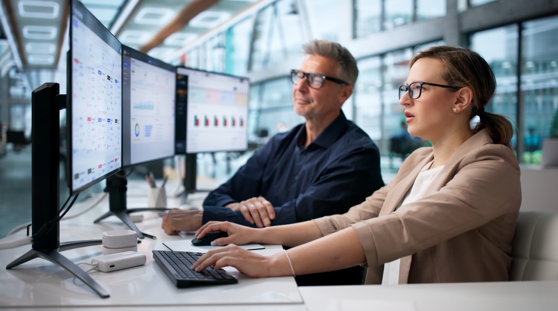 Two IT specialists reviewing monitoring dashboards across three screens in a bright office.