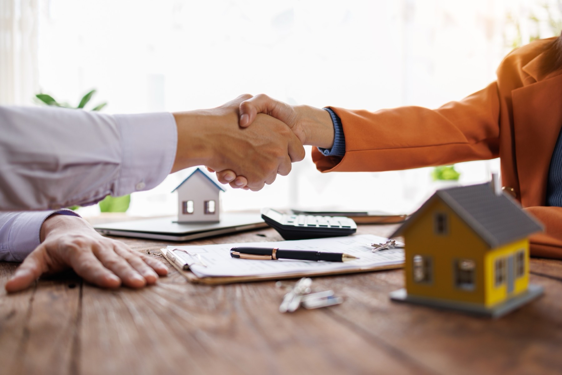 Two people shaking hands over a signed property agreement, with model buildings, keys and a calculator on the desk